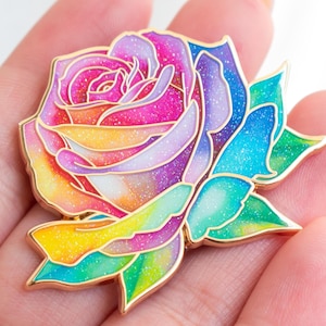 May include: A rose-shaped enamel pin featuring a vibrant rainbow gradient of pink, purple, blue, yellow, and green colors. The design includes gold-toned metal outlines separating each petal, and the entire surface has a fine, shimmering glitter finish.