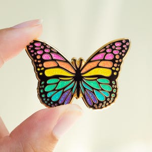 May include: A colorful butterfly enamel pin with a gold outline. The butterfly's wings feature a rainbow of colors, including pink, orange, yellow, green, and purple. The pin is held between two fingers.