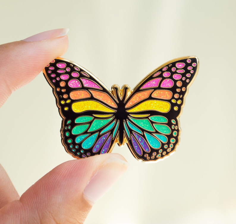 May include: A colorful butterfly enamel pin with a gold outline. The butterfly's wings feature a rainbow of colors, including pink, orange, yellow, green, and purple. The pin is held between two fingers.