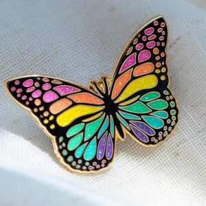May include: A colorful butterfly enamel pin with a gold outline. The butterfly's wings are black with sections of pink, orange, yellow, green, and blue. The pin is approximately 2 inches wide and is set against a white background.