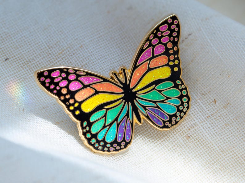 May include: A colorful butterfly enamel pin with a gold outline. The butterfly's wings are black with sections of pink, orange, yellow, green, and blue. The pin is approximately 2 inches wide and is set against a white background.