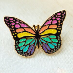 May include: A colorful butterfly pin with a gold outline. The wings are filled with vibrant colors including pink, orange, yellow, green, and purple. The pin is a decorative accessory.