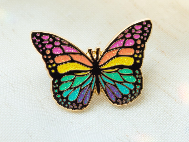 May include: A colorful butterfly pin with a gold outline. The wings are filled with vibrant colors including pink, orange, yellow, green, and purple. The pin is a decorative accessory.