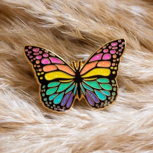 May include: A colorful butterfly pin with a gold outline. The wings feature a gradient of pink, orange, yellow, green, and blue. The pin is set against a background of fluffy, light brown material.