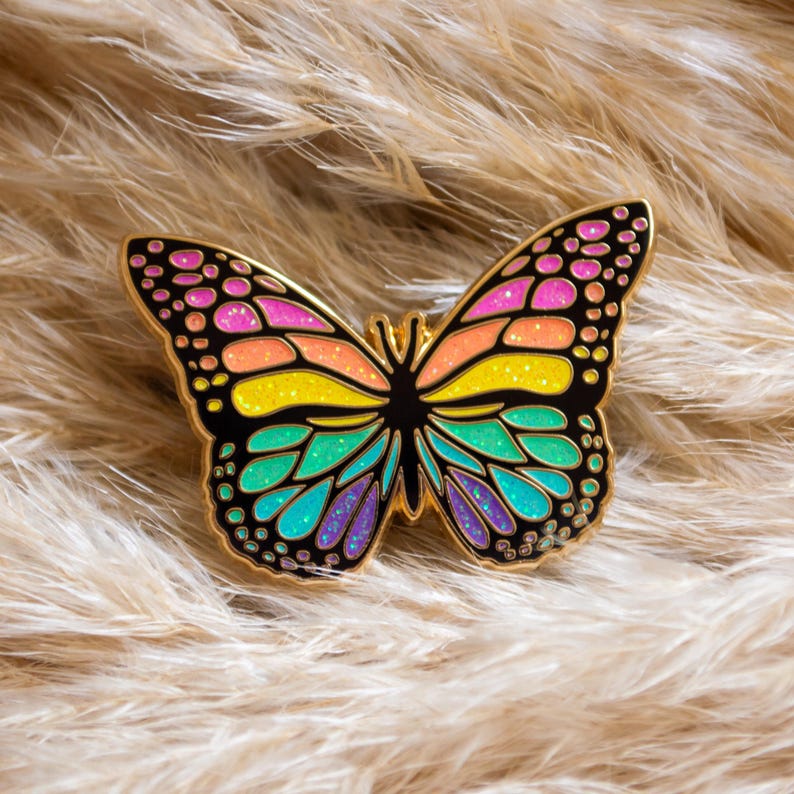 May include: A colorful butterfly pin with a gold outline. The wings feature a gradient of pink, orange, yellow, green, and blue. The pin is set against a background of fluffy, light brown material.