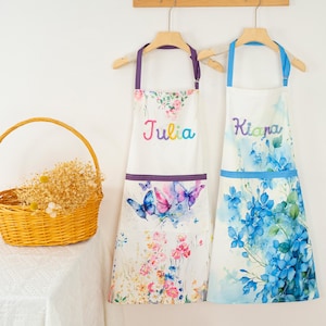 May include: Two white aprons with floral and butterfly designs hang on a wooden rack. One apron has the name "Julia" in colorful script, and the other has "Kiana". A yellow wicker basket filled with dried flowers sits on a white surface.