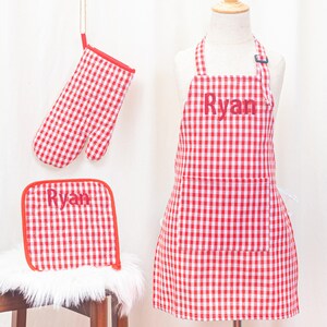 May include: A red and white checkered apron, oven mitt, and pot holder set. The apron has a pocket and the name "Ryan" printed on it. The oven mitt and pot holder also have the name "Ryan" printed on them. The set is displayed on a white background.