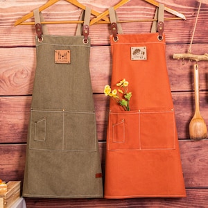 May include: Two aprons hanging on wooden hangers. One is olive green, the other is orange. Both have pockets, leather accents, and adjustable straps. The olive green apron has the name "Charlie" on it. The orange apron has the name "Tony" on it.