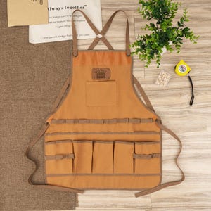 May include: A tan canvas apron with multiple pockets, ideal for gardening or crafting. Features a leather patch with a crown logo and adjustable straps. The apron is designed for functionality and durability, perfect for protecting clothing while working on projects.