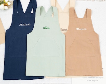 Puede incluir: Cuatro delantales de lino personalizados en diferentes colores. Los delantales son azul marino, verde claro, beige y marrón. Cada delantal tiene un bolsillo y correas ajustables. Los nombres Adelaide, Ava, Emma y Monica están bordados en los delantales.