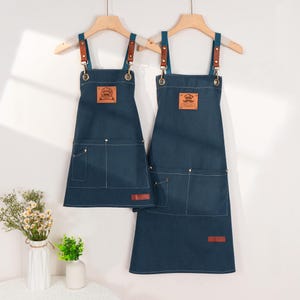 May include: Two dark blue denim aprons suspended from wooden hangers. Each apron features brown leather straps and a front pocket. One apron displays a chef's hat logo, the other a moustache logo. Both have a small brown leather tag.