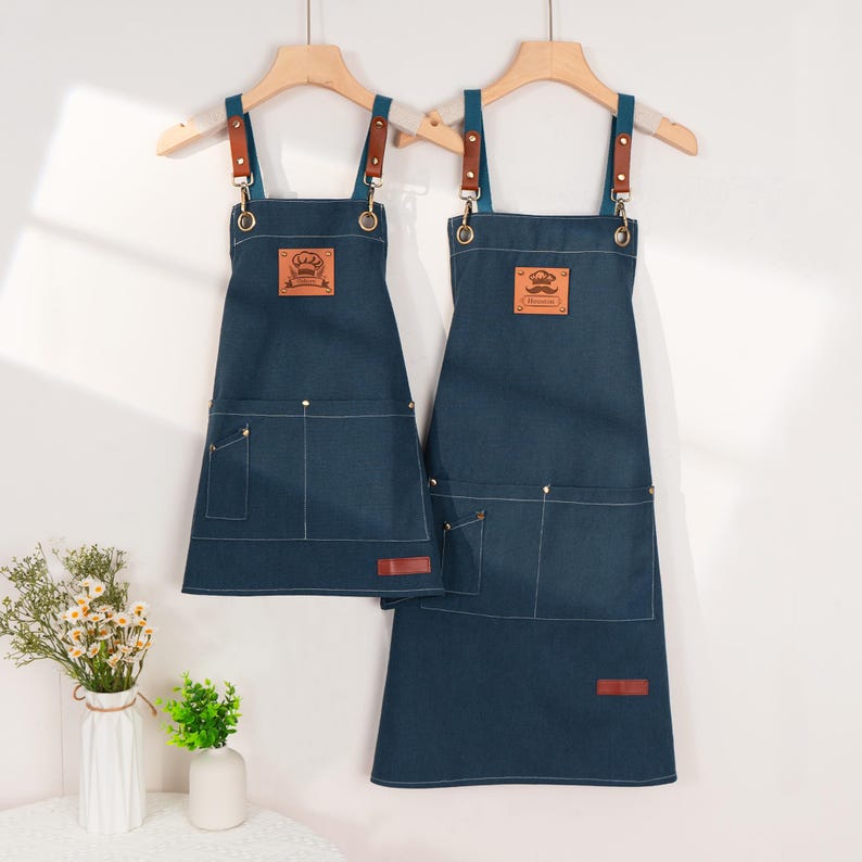 May include: Two dark blue denim aprons suspended from wooden hangers. Each apron features brown leather straps and a front pocket. One apron displays a chef's hat logo, the other a moustache logo. Both have a small brown leather tag.