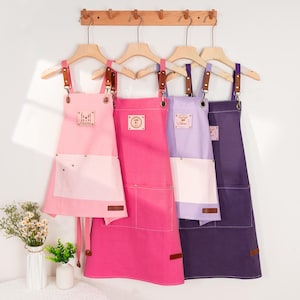 May include: Four aprons in shades of pink, purple, and indigo hang on wooden hangers. Each apron has a contrasting white pocket and leather accents. The aprons have text that reads "Charlie", "Cara", and "Oliver".