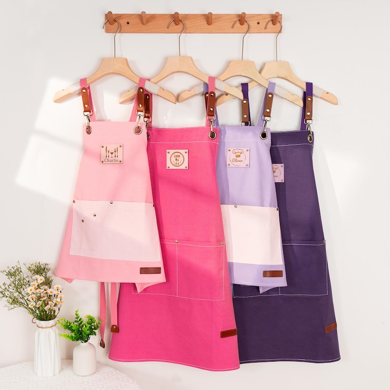 May include: Four aprons in shades of pink, purple, and indigo hang on wooden hangers. Each apron has a contrasting white pocket and leather accents. The aprons have text that reads "Charlie", "Cara", and "Oliver".