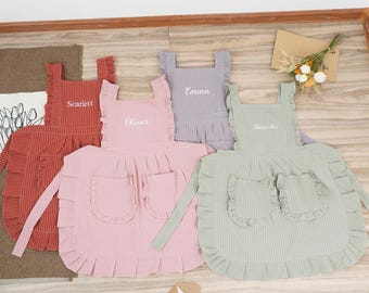 Op de afbeelding: Vier gepersonaliseerde kinder-schorten in roze, groen, paars en rood met witte strepen. Elke schort heeft een ruche-bies en twee zakken. De schorten zijn geborduurd met de namen "Oliver", "Emma", "Natasha" en "Scarlett".