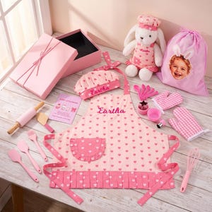 May include: A pink baking set featuring an apron and chef hat embroidered with the name Eartha, a plush bunny, a drawstring bag with a child's photo, a rolling pin, spatulas, a whisk, oven mitts, and cookie cutters in a pink gift box.