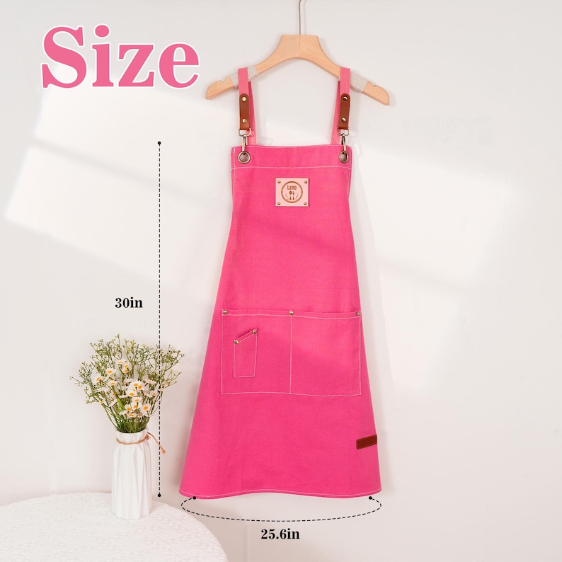 May include: A vibrant pink apron with brown leather straps and a wooden hanger. The apron features two front pockets and a small patch with text. The apron is 76 cm long and 65 cm wide.