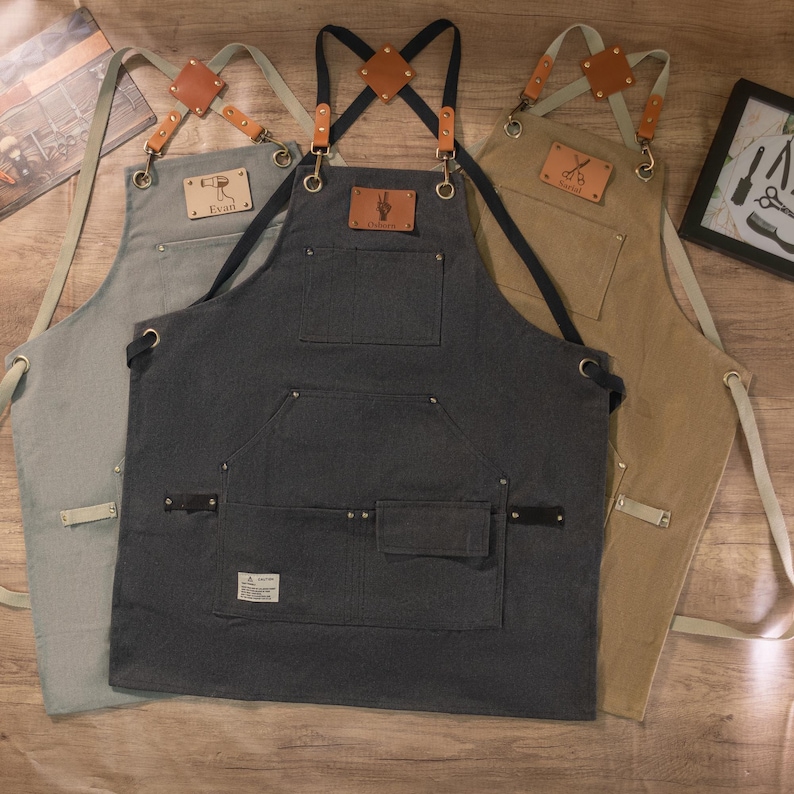 Op de afbeelding: Drie schorten van canvas met verstelbare banden en leren accenten. De schorten zijn in verschillende kleuren: lichtblauw, donkergrijs en bruin. Elke schort heeft een leren patch met een ander ontwerp en tekst. De tekst op de patches luidt "Evan", "Barber" en "Scissors".