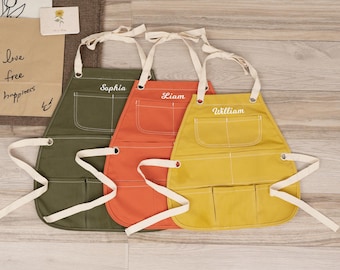 Puede incluir: Tres delantales de lona personalizados con correas ajustables y múltiples bolsillos. Los delantales son de color verde, naranja y amarillo con los nombres "Sophia", "Liam" y "William" bordados en blanco.