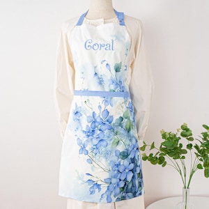 May include: White apron with a blue floral design and the word "Coral" in script. The apron features a light blue trim and straps. The floral pattern includes blue flowers and green leaves, creating a watercolor effect.