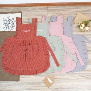 May include: Four striped aprons with ruffled edges in red, green, pink, and gray. Each apron has two pockets and adjustable straps. The aprons are personalized with the names "Scarlett" and "Natasha".