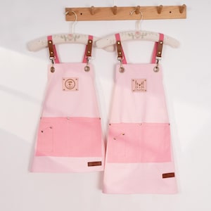 May include: Two pink aprons with darker pink pockets and accents. The aprons have brown leather straps and are hanging on decorative hangers. One apron has the word "Ludo" and the other has the word "Charlie" on a small patch.