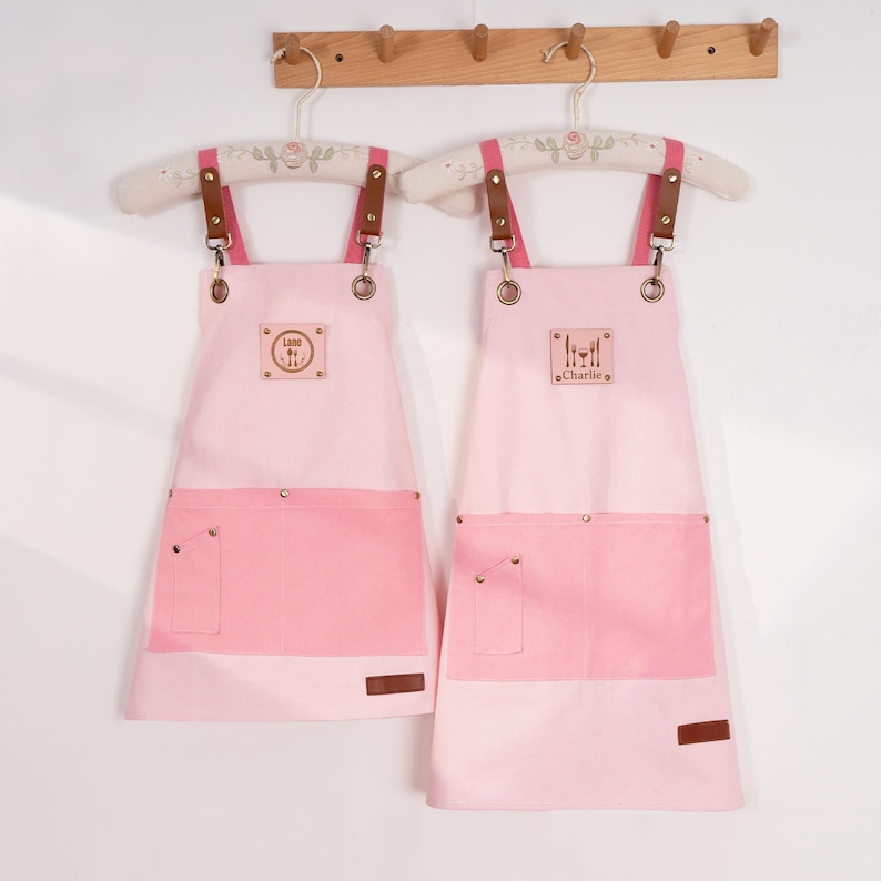 May include: Two pink aprons with darker pink pockets and accents. The aprons have brown leather straps and are hanging on decorative hangers. One apron has the word "Ludo" and the other has the word "Charlie" on a small patch.