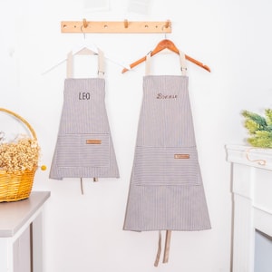 May include: Two striped aprons hanging on a wall. One apron is smaller and says "LEO", the other is larger and says "Bonnie". Both aprons have a pocket and a leather patch.