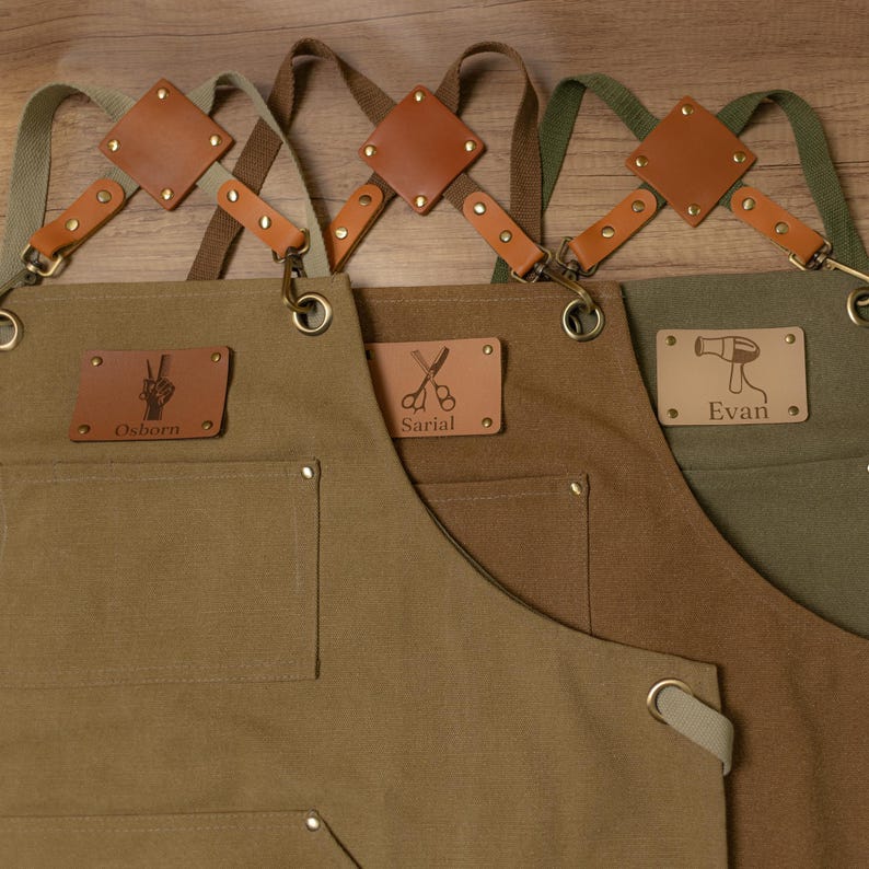 Op de afbeelding: Drie bruine canvas schorten met verstelbare banden en leren accenten. Elke schort heeft een gepersonaliseerde leren patch met een naam en een symbool: een vredesteken, een schaar en een f&ouml;hn.