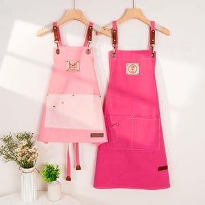 May include: Two pink aprons hanging on wooden hangers. The aprons have brown leather straps and pockets. One apron is light pink with a white pocket and the name "Charlie". The other apron is a darker pink.