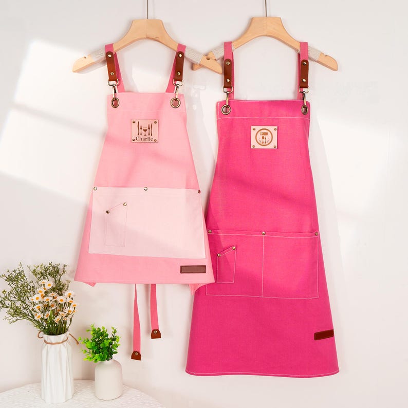 May include: Two pink aprons hanging on wooden hangers. The aprons have brown leather straps and pockets. One apron is light pink with a white pocket and the name "Charlie". The other apron is a darker pink.