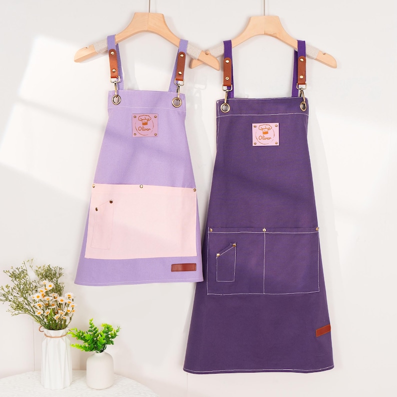 May include: Two aprons hanging on wooden hangers. One is lavender with a pink pocket, and the other is purple. Both have brown leather straps and a small patch with a chef's hat logo and the name "Oliver".