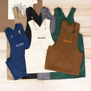 May include: A collection of personalized aprons in various colors, including navy, tan, black, cream, gray, and brown. Each apron features a large front pocket and cross-back straps. Some aprons are embroidered with names like "Kinley," "Blakely," and "Charlotte."