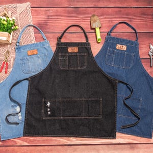 May include: Three denim aprons in varying shades of blue and black. Each apron has a large front pocket, a leather patch, and a neck loop. The aprons are designed for kitchen or workshop use. The background is a wooden surface.