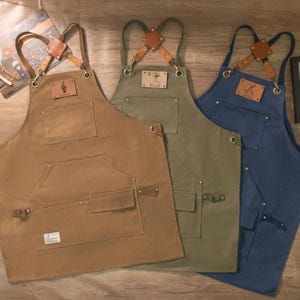 Op de afbeelding: Drie schorten van canvas met verstelbare banden en leren accenten. De schorten zijn bruin, groen en blauw. Elke schort heeft een leren patch met een ander ontwerp en de naam "Evan" of "Barber".