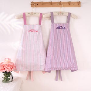 May include: Two aprons hanging on a wooden rack. One is pink and white striped with the name "Alice" embroidered in pink. The other is purple and white striped with "Bliss" embroidered in purple. Both have matching straps and ties.