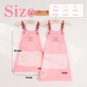 May include: Two pink aprons with white pockets and brown leather straps hang on a wooden rack. The aprons have a small logo patch and are labeled with size measurements: width 50cm and 53cm, height 60cm and 63cm.