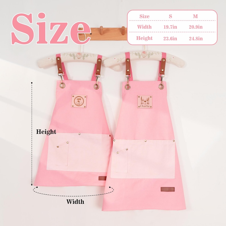 May include: Two pink aprons with white pockets and brown leather straps hang on a wooden rack. The aprons have a small logo patch and are labeled with size measurements: width 50cm and 53cm, height 60cm and 63cm.
