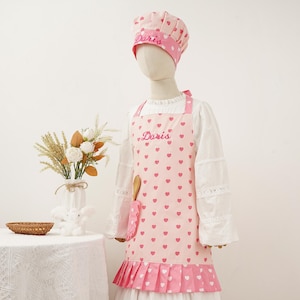 May include: A pink apron and chef's hat set with a heart pattern. The apron has the name "Doris" embroidered on it and a pocket for a wooden spoon. The hat also has the name "Doris" embroidered on it.