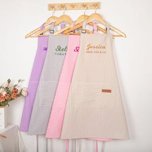 May include: Four striped aprons in purple, gray, pink, and beige hang on wooden hangers. Each apron has a pocket and personalized text: "M", "Stella Cookie", "Si", and "Jessica Made with Love".