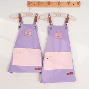 May include: Two lavender aprons with light pink pockets and brown leather straps hang from a wooden rack. Each apron has a small square patch with a chef's hat logo and the name "Oliver". The aprons are designed for kitchen use.
