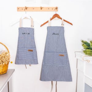 May include: Two blue and white pinstriped aprons hanging on wooden hangers. Each apron features a front pocket with a small brown rectangular patch and embroidered text on the chest, reading Sawyer on the smaller one and Mason on the larger one.