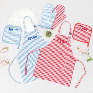 May include: A set of personalized kitchen accessories, including a blue and white gingham apron, pot holder, and oven mitts, with the name "Jason" embroidered on the apron and pot holder. A red and white gingham apron, pot holder, and oven mitts with the name "Ryan".