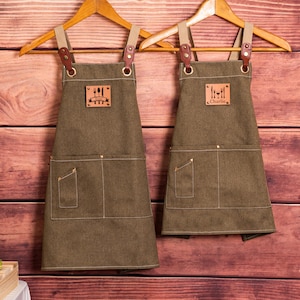 May include: Two olive green aprons with brown leather straps and pockets, hanging on wooden hangers. Each apron has a leather patch with a utensil design. One apron is personalized with the name "Charlie".