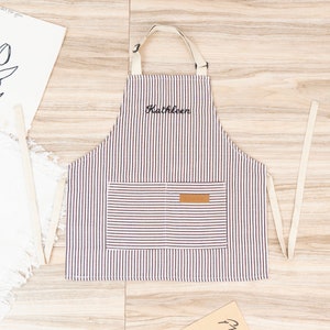 May include: A white and brown striped kitchen apron with a brown pocket and a brown leather label that says "EDOK" on it. The apron has adjustable straps and the name "Kathleen" is embroidered on the front.