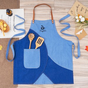 May include: A two-tone denim apron with a brown leather neck strap. The apron has a pocket and includes wooden cooking utensils. The apron is personalized with the name "Taylor" and a coffee cup design.