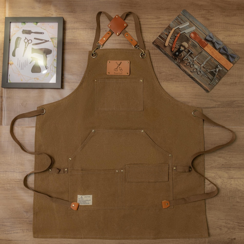 Op de afbeelding: Een bruine canvas schort met verstelbare banden en meerdere zakken. Het schort heeft een leren patch met het woord "Barbers" en een gekruist schaar.