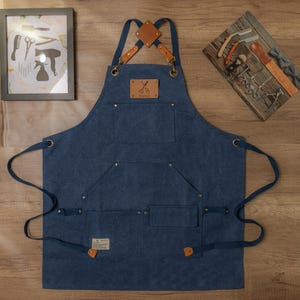 Op de afbeelding: Een blauwe denim schort met bruine leren banden en een bruine leren patch met een schaar en het woord "Barbers". Het schort heeft meerdere zakken en is ontworpen voor een barbier.