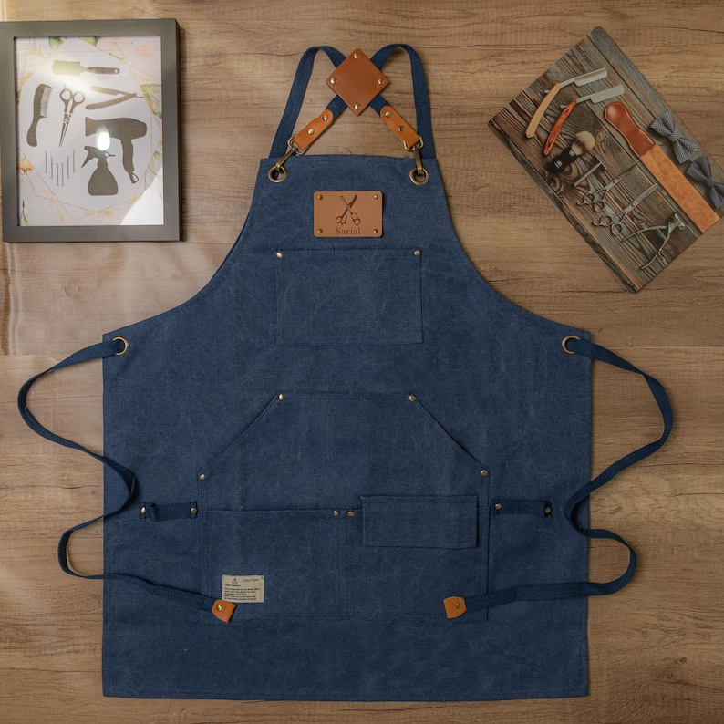 Op de afbeelding: Een blauwe denim schort met bruine leren banden en een bruine leren patch met een schaar en het woord "Barbers". Het schort heeft meerdere zakken en is ontworpen voor een barbier.