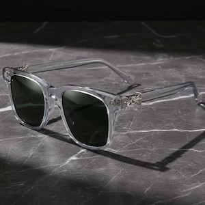 May include: Transparent-framed sunglasses with dark lenses. The square frames feature decorative silver detailing at the temples. The sunglasses are set against a dark, marbled surface.